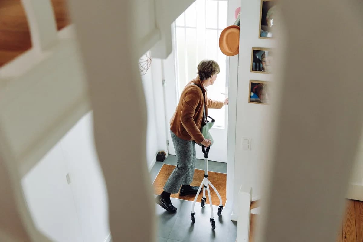 An elderly woman uses a walker at the doorway of a home.