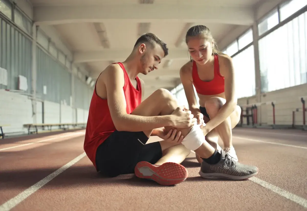 A woman tapes a man's injured leg while they sit on an indoor track.