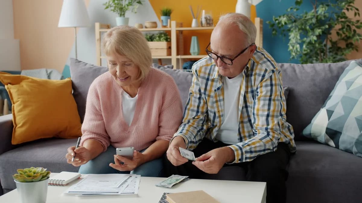 An elderly couple sits on a couch reviewing bills and cash at home.
