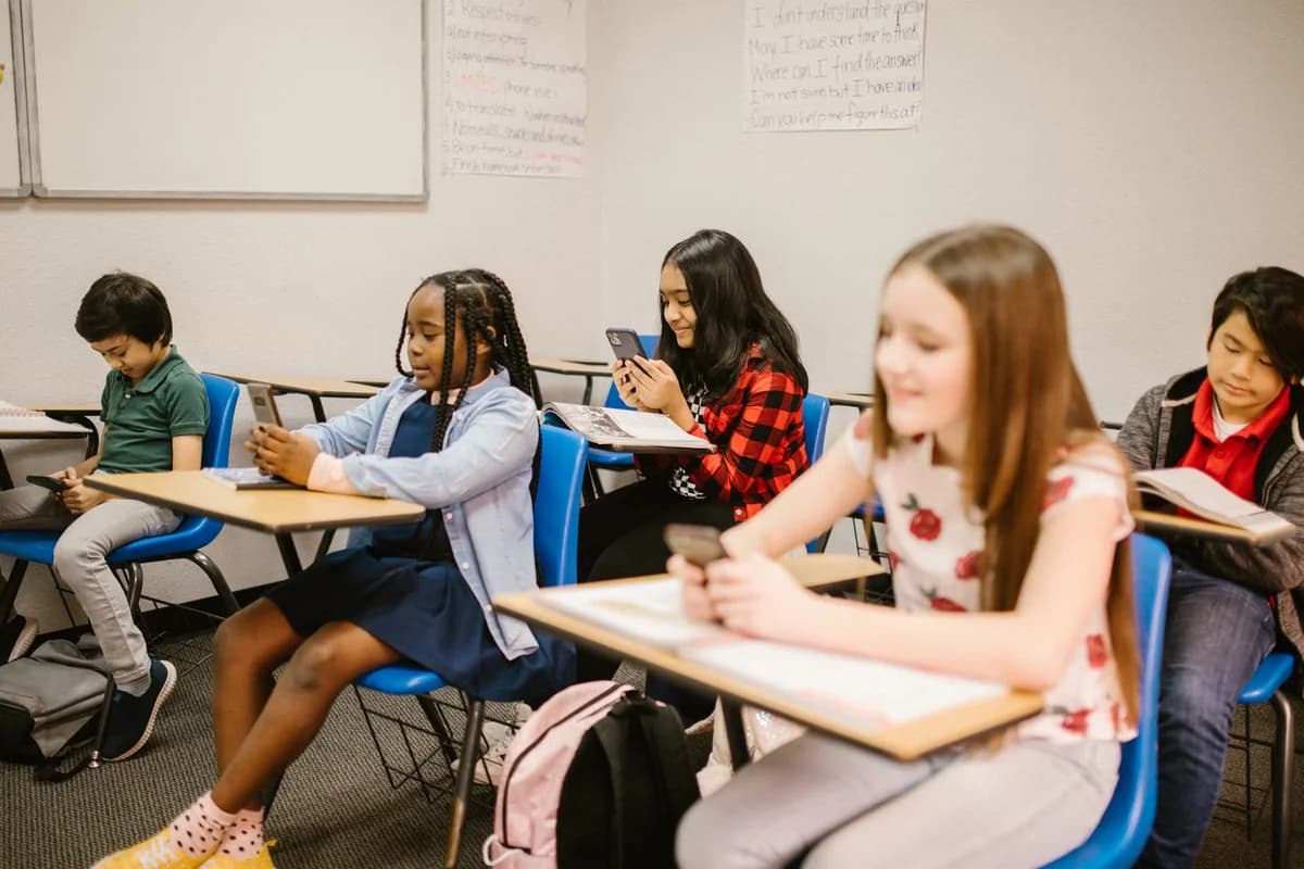 Schoolchildren sitting at desks in a classroom, using mobile phones.