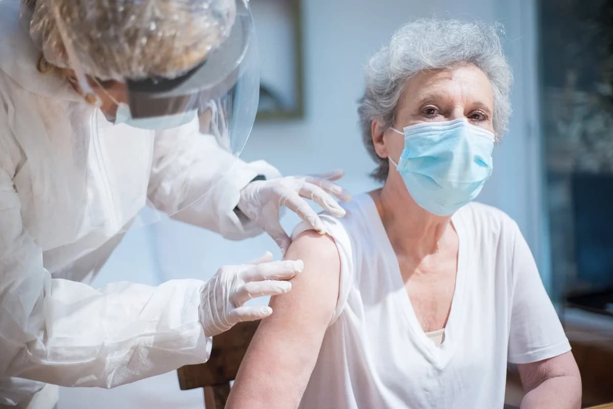 An older masked woman is vaccinated by a healthcare worker in PPE.