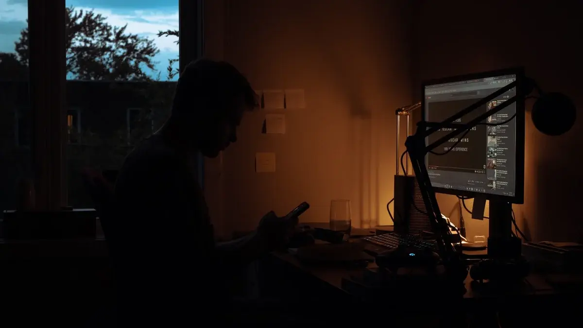 Silhouette of a man using a smartphone beside a glowing computer monitor in a dark room.