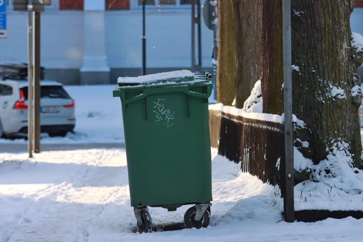 A green trash bin covered with snow stands on a snowy sidewalk next to a tree and fence.