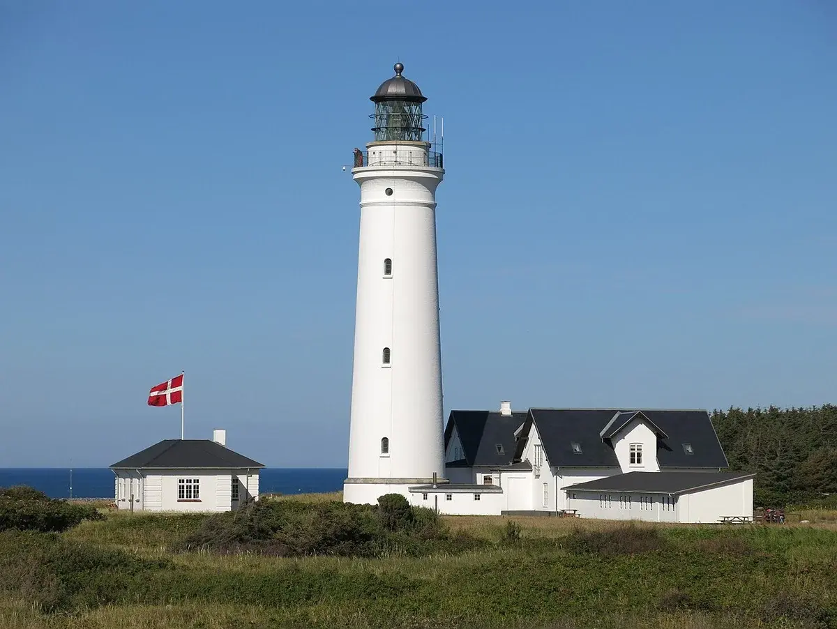 A tall white lighthouse by the coast with nearby buildings and a Danish flag.