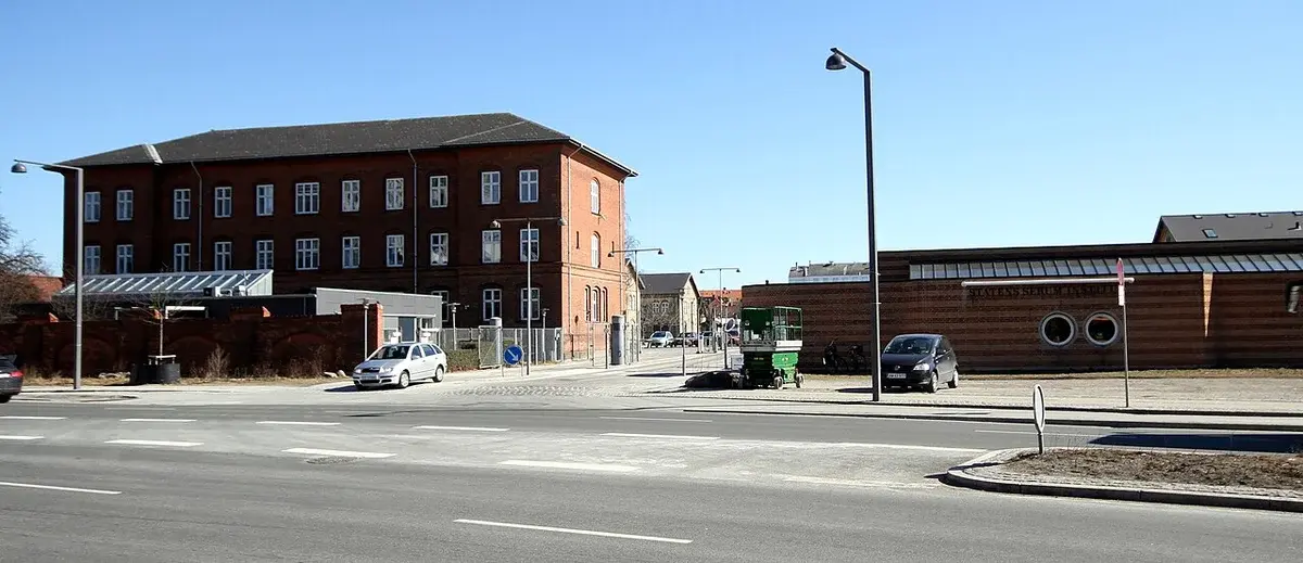 Street view of the Statens Serum Institut entrance with brick buildings and cars in Copenhagen.