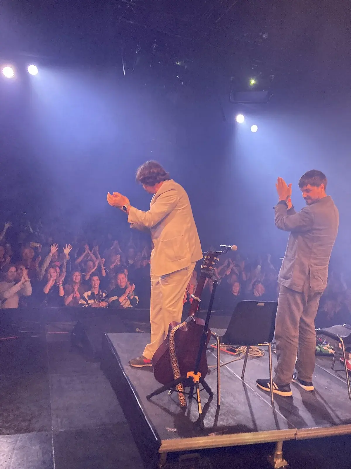 Two performers clapping onstage at a concert with a crowd in front.