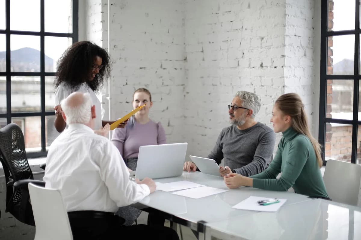 A Black woman presents to coworkers seated around a table in a bright office.