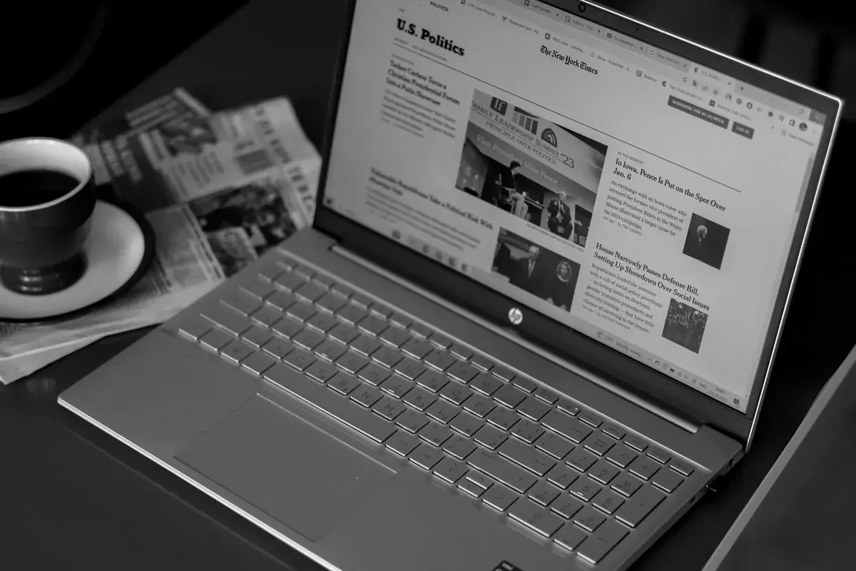 A black-and-white photo of a laptop showing news articles beside coffee and newspapers.