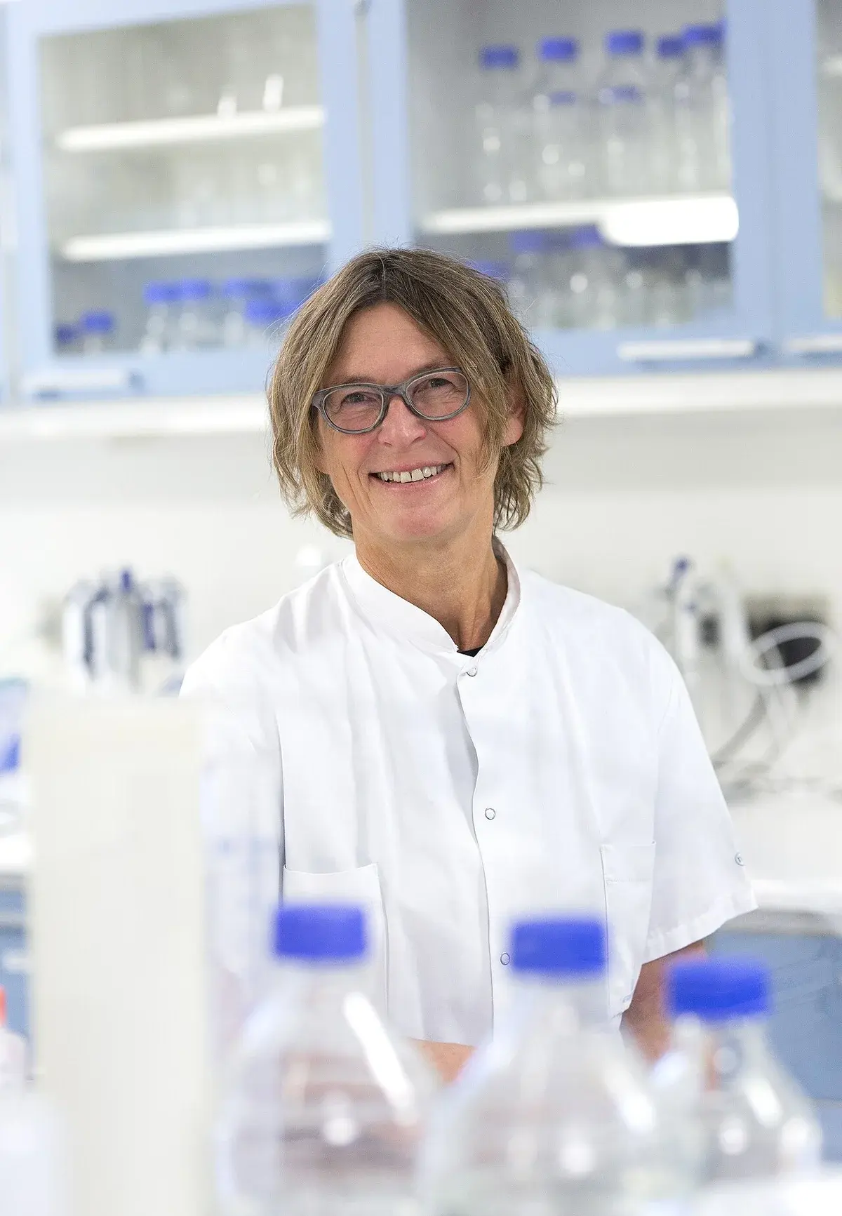 A smiling woman in a white lab coat standing in a laboratory.