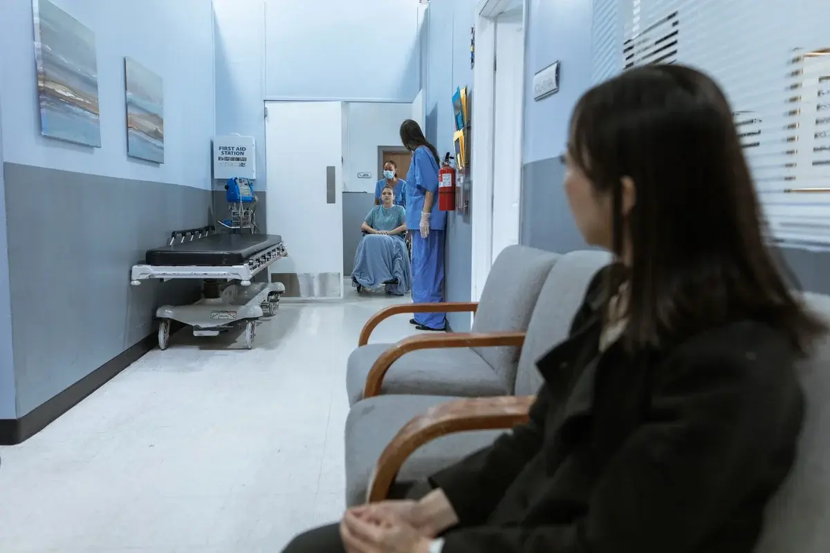 A hospital hallway with a patient in a wheelchair and medical staff nearby, with a seated woman in the foreground.