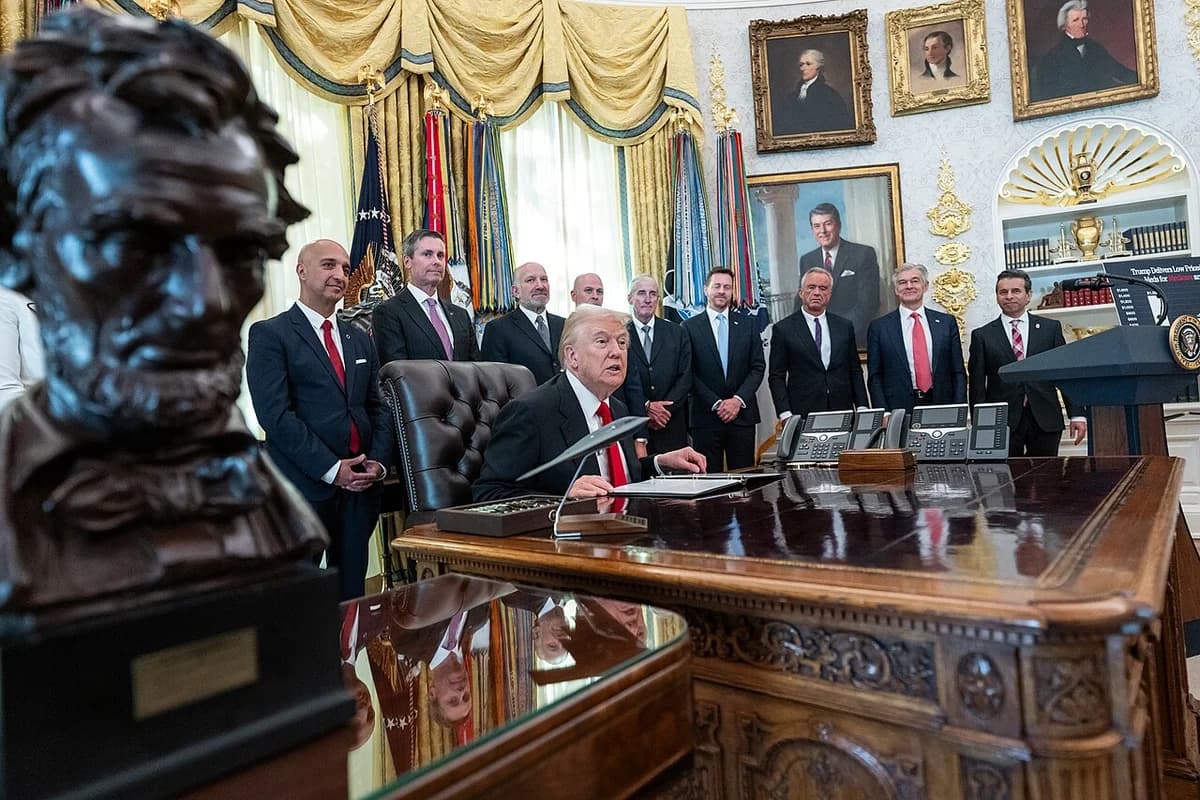Donald Trump seated at the Resolute Desk in the Oval Office with several men standing behind him.