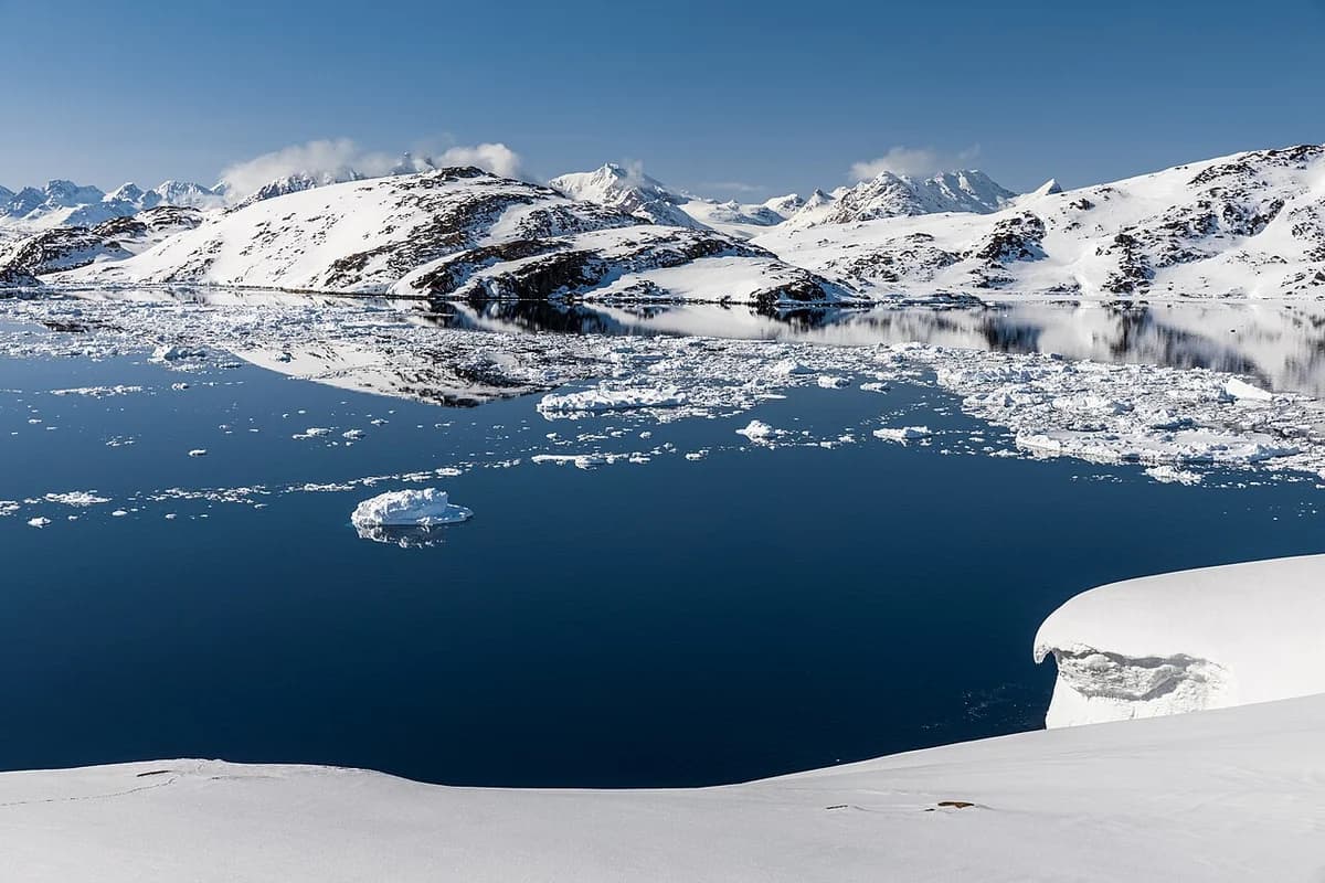 Snow-covered mountains reflected in calm Arctic water with floating ice.