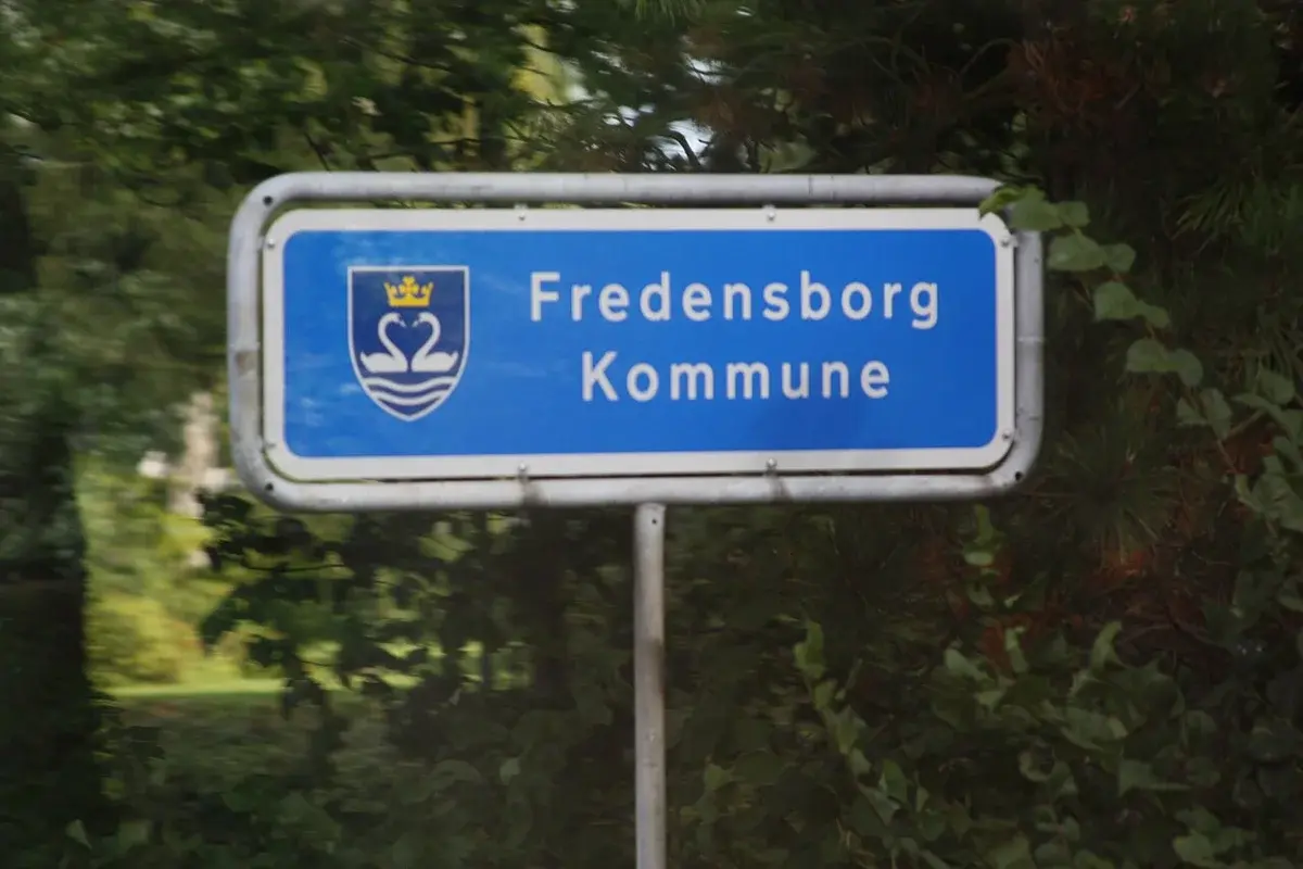 Blue sign reading Fredensborg Kommune beside trees and bushes.